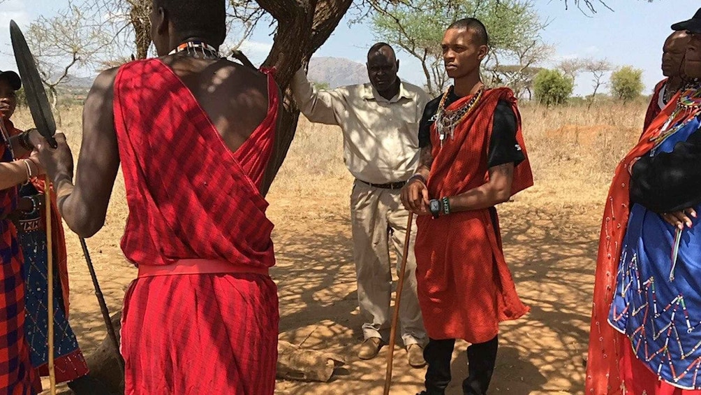 A group of people in traditional attire, standing in a dry, open landscape with sparse trees. 4