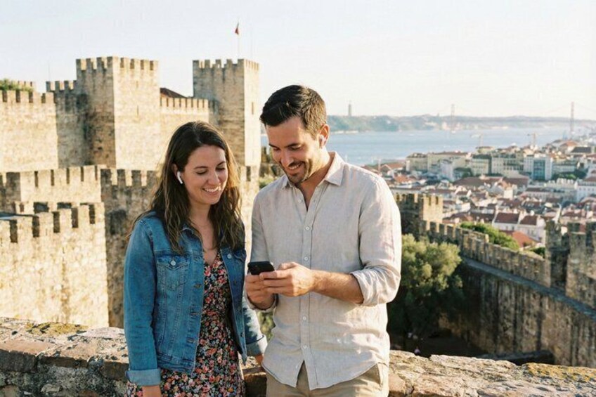 Two people standing on a stone wall overlooking a city with historic buildings and a castle in the background. 4