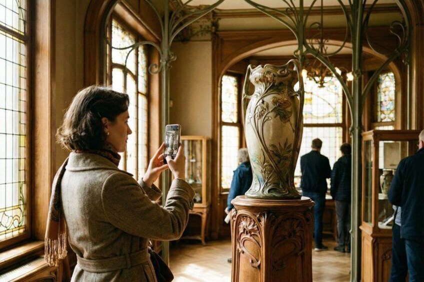 A person taking a photo of a large ornate vase in a museum with stained glass windows and visitors in the background. 3