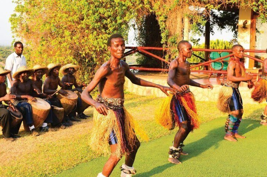A group of people in traditional attire performing a dance, with others seated and playing drums in the background. 3