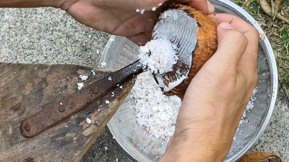 Hands using a knife to cut a coconut, with the coconut pieces and white flesh falling into a metal bowl. 2