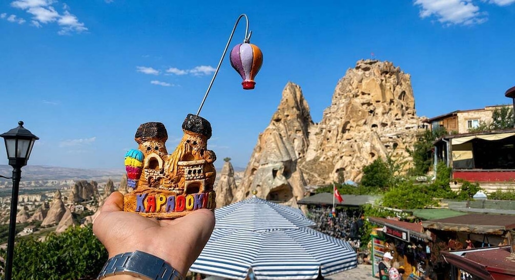 A hand holding a hot air balloon toy with a string, a rocky mountain backdrop, and a crowd of people in the foreground. 3