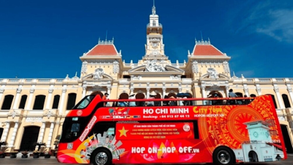 A red double-decker bus with 'HO CHI MINH CITY TOUR' written on it, parked in front of a large, ornate building with two towers. 2