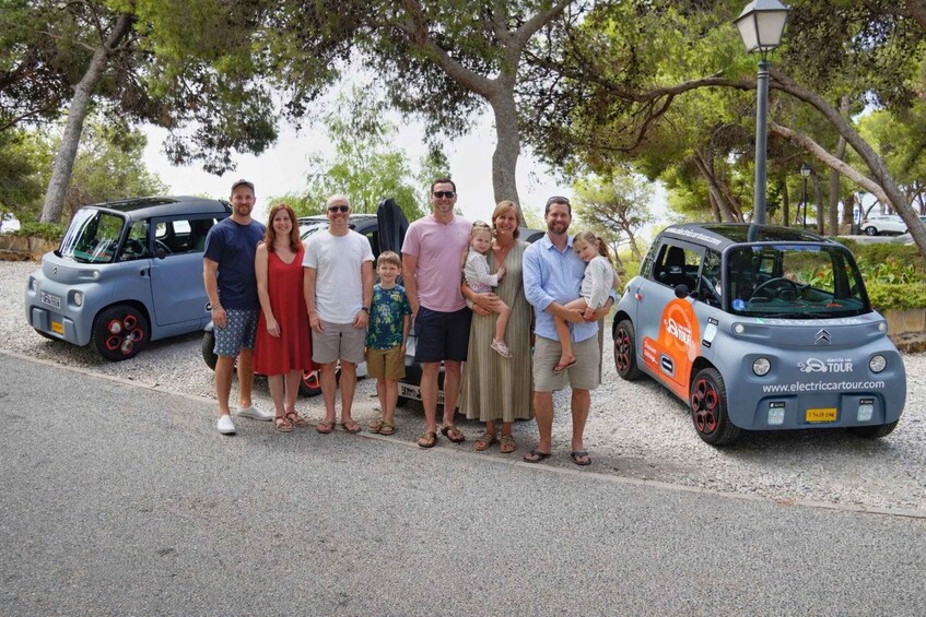 A group of people standing in front of a small electric car and a blue car on a road with trees in the background. 2