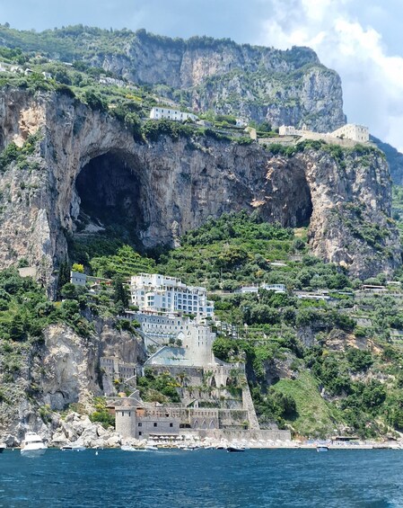 A coastal town with buildings on a cliff, a large cave-like opening, and boats in the water. 3