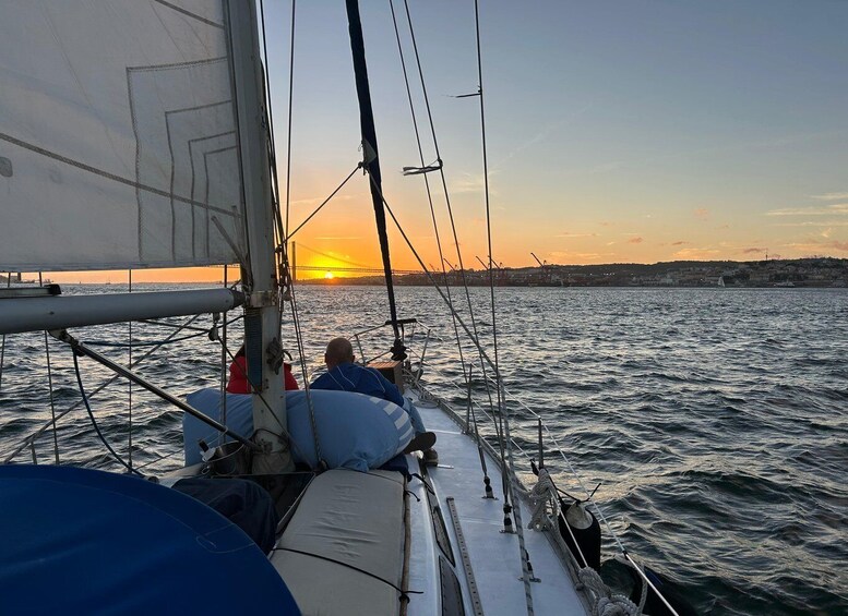 A sailboat with two people on deck, a sunset in the background, and a bridge across the water. 5