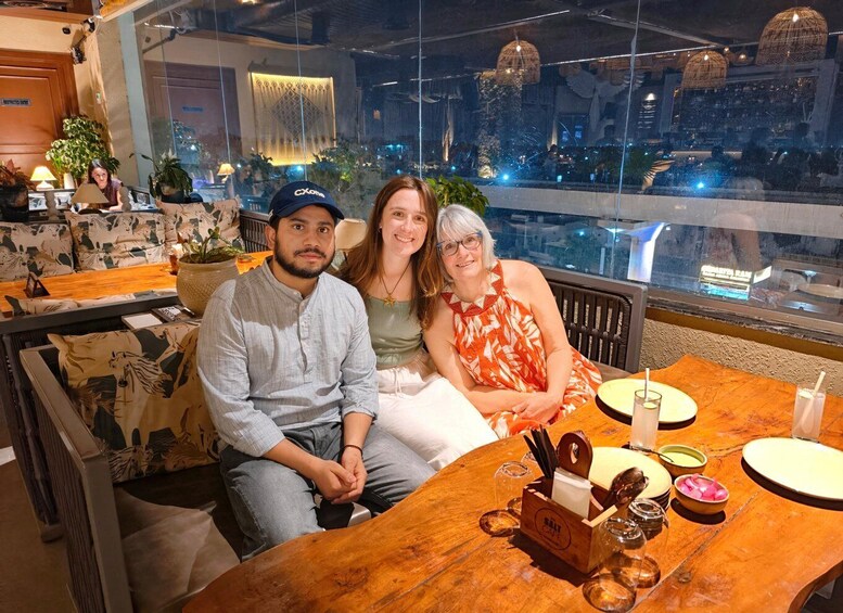 Three people seated at a wooden table in a restaurant with a view of the cityscape. 2