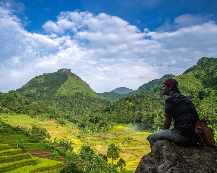 A person sitting on a rock overlooking terraced fields and a hill with a castle-like structure. 2