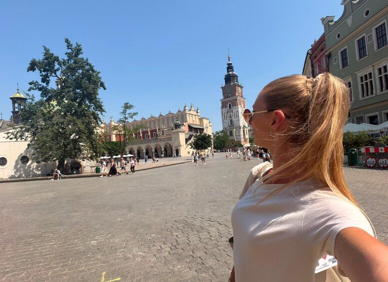 A woman taking a selfie in a public square with historic buildings and a clock tower in the background. 1