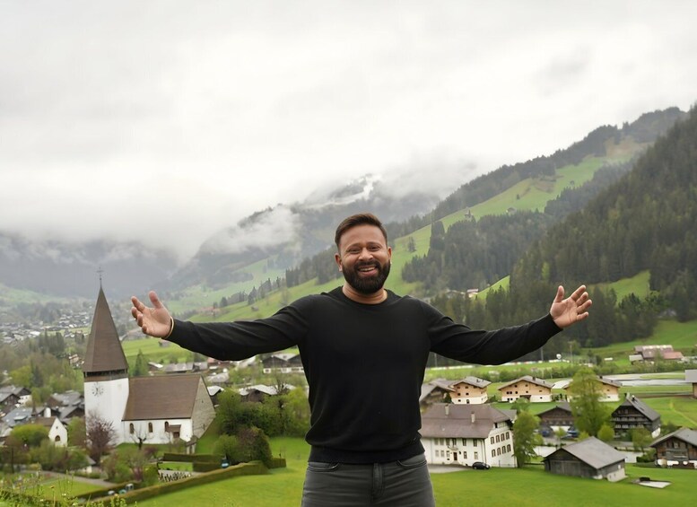 A person standing with arms outstretched in a mountainous area with a church and houses in the background. 5