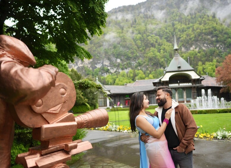 A couple embracing near a large bronze sculpture in a park with a traditional building in the background. 3