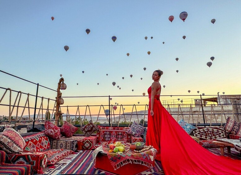 A woman in a red dress standing on a rooftop with a view of hot air balloons in the sky. 1