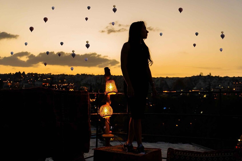 A silhouette of a person standing on a rooftop with a cityscape in the background and hot air balloons in the sky. 4