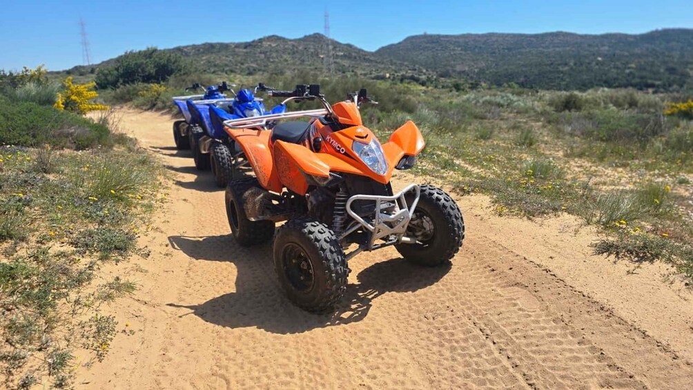 Two quad bikes on a dirt path with sparse vegetation and hills in the background. 3