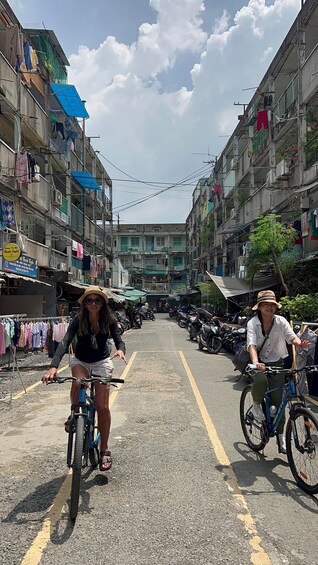 Two people riding bicycles down a street lined with multi-story buildings, some with balconies and laundry hanging out to dry. 3