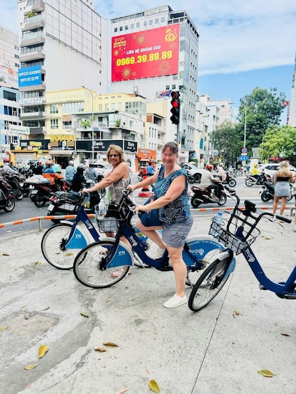 Two women riding electric bikes on a busy street with buildings and shops in the background. 5