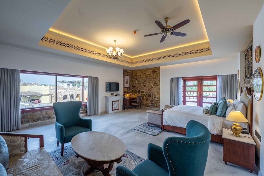 A hotel room with a bed, two armchairs, a round table, a ceiling fan, a TV, and a stone wall. 2