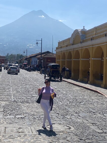 Una calle empedrada con una mujer caminando, un edificio amarillo con arcos y una montaña al fondo. 3
