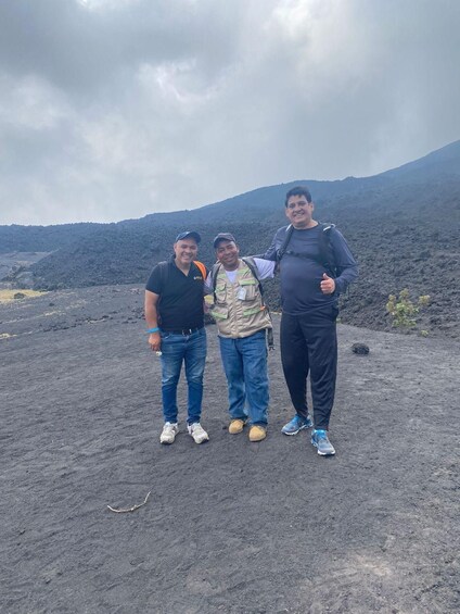 Tres personas de pie en un paisaje volcánico con una montaña al fondo. 1