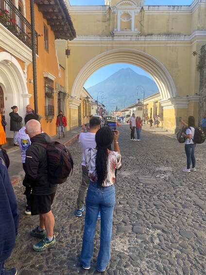Una calle adoquinada con gente fotografiando, una montaña al fondo y edificios históricos con arcos. 2