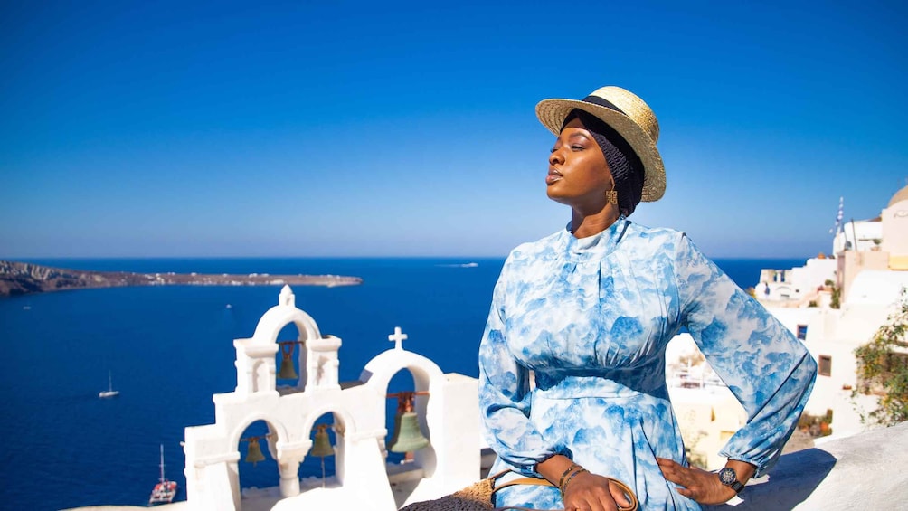 A person in a blue patterned dress and straw hat overlooking a coastal town with white buildings and a blue sea. 1
