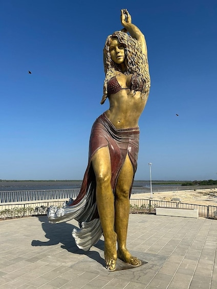 A bronze statue of a woman in a flowing dress, standing on a paved area with a beach and fence in the background. 4
