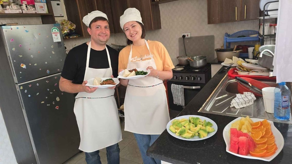 Two people in a kitchen holding plates of food, with a stove, sink, and various kitchen items in the background. 3
