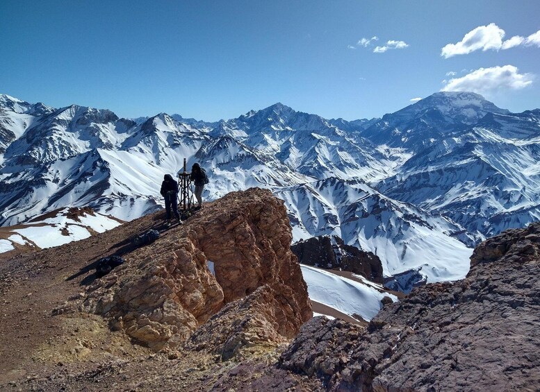 A mountainous landscape with snow-covered peaks and a rocky outcrop where two hikers are standing. 3