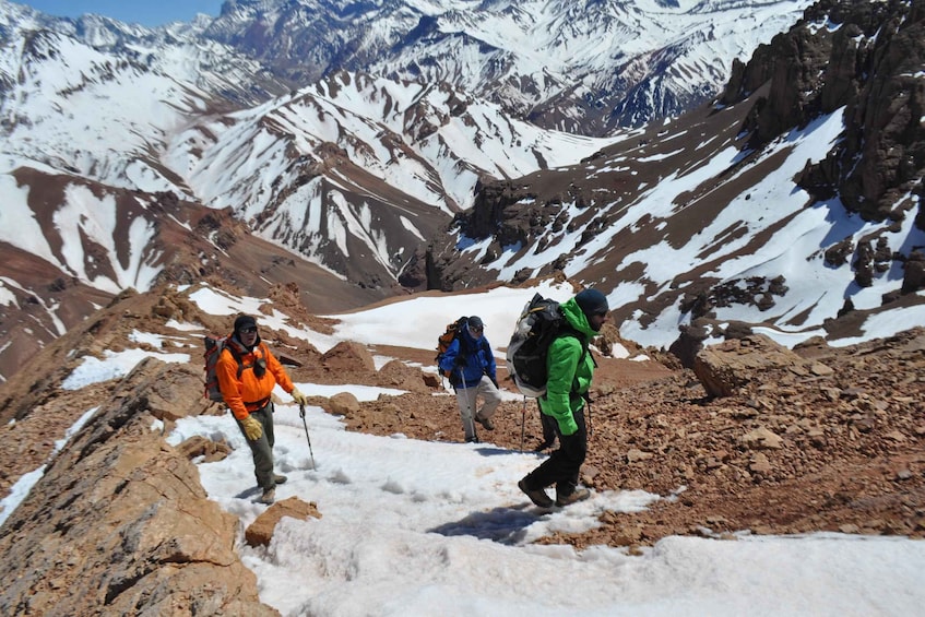 Three hikers on a rocky mountain trail with snow patches and snow-capped mountains in the background. 1