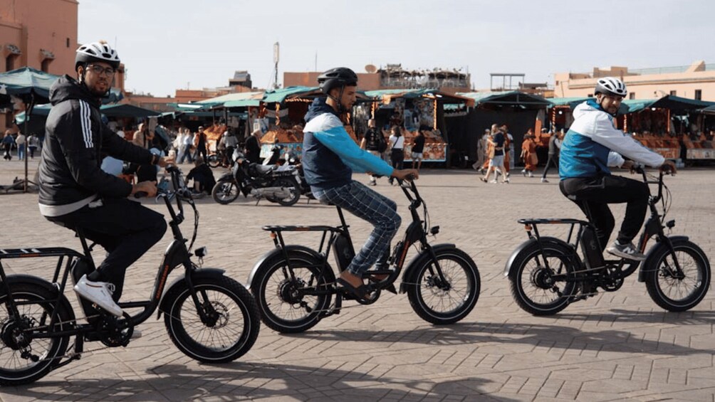 Three people riding electric bikes in a busy outdoor market area. 1