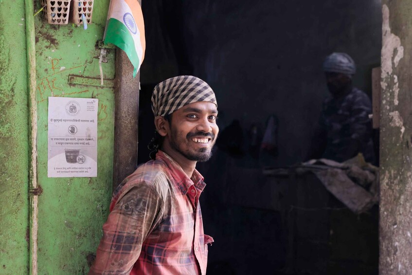 A man in a plaid shirt standing in front of a green door with an Indian flag and a sign. 3