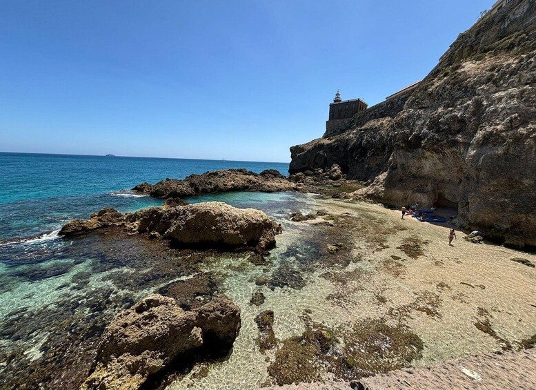 A rocky coastline with clear water, a lighthouse, and people on the beach. 4
