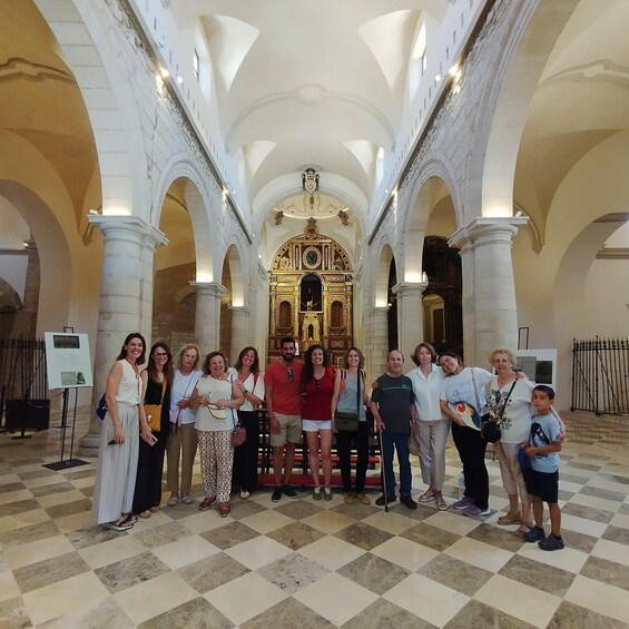A group of people standing in a grand hall with high ceilings and arches, a central ornate altar, and a checkered floor. 3