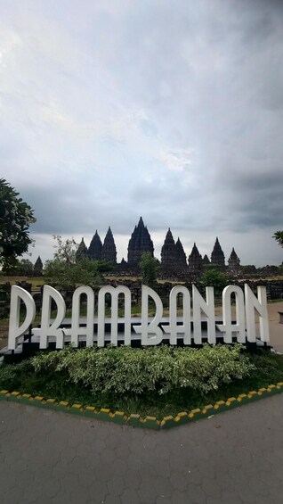 A sign with 'PRAMBANAN' in large letters, surrounded by greenery and a temple complex in the background. 3