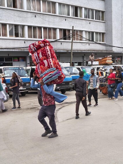 A street scene with a man carrying a large, colorful fabric on his head, people walking, and parked cars. 4