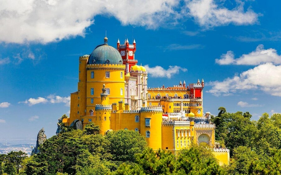 A yellow castle with a red tower and a clock, surrounded by greenery. 1