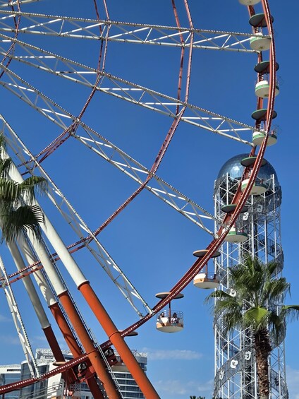 A Ferris wheel with red and white gondolas, a palm tree, and a tall tower with a glass facade. 1