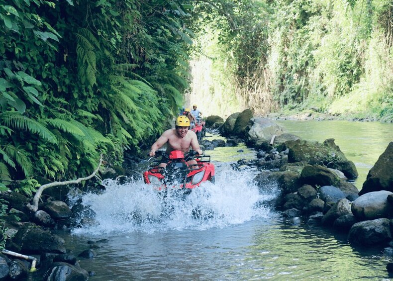 A person riding a red ATV through a shallow stream surrounded by dense green foliage. 3