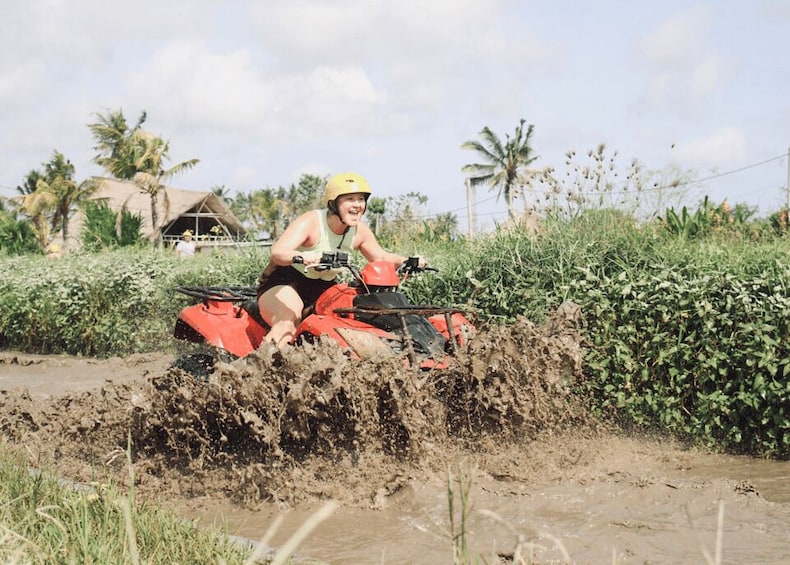 A person riding a red ATV through a muddy area with vegetation and a house in the background. 5
