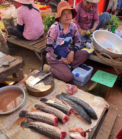 A market scene with people sitting on benches, a woman cutting fish, and various food items displayed. 3