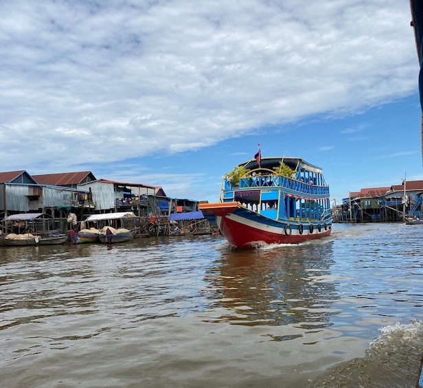 A colorful boat with a blue and red hull navigating through a river with floating houses. 2