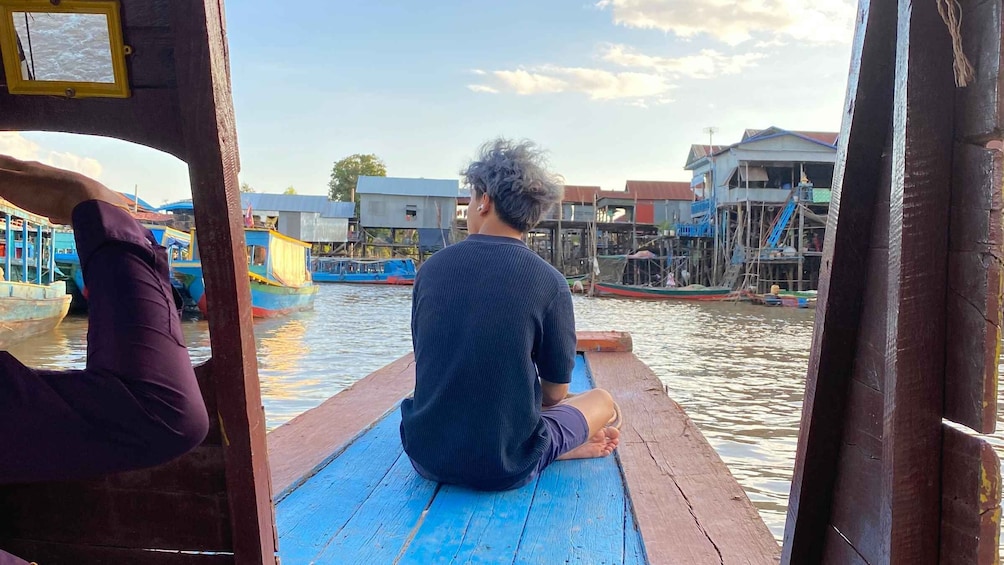 A person sitting on a wooden dock overlooking a river with boats and houses. 1