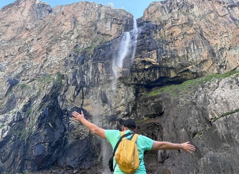 A person with arms outstretched facing a waterfall cascading down a rocky cliff. 1