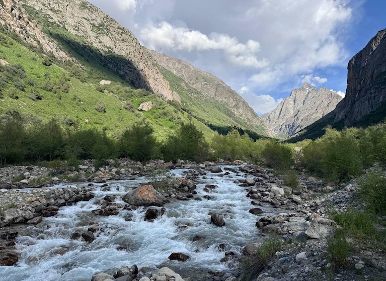 A fast-flowing river with rocky bed, surrounded by green hills and mountains under a partly cloudy sky. 2