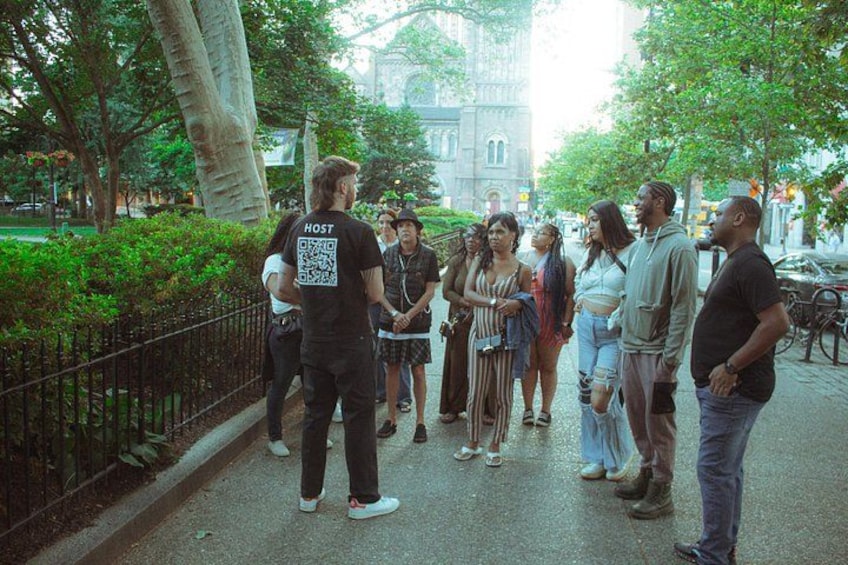 A group of people standing on a sidewalk, a large tree, and a building in the background. 3