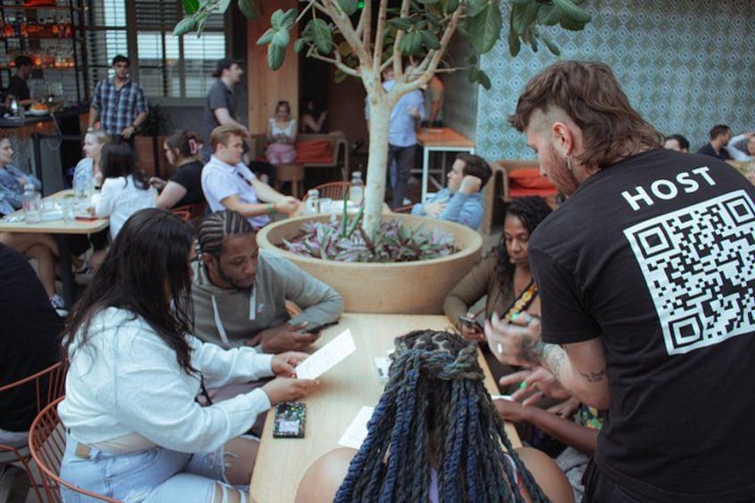A casual dining setting with people seated at tables, a potted plant, and a QR code on a person's shirt. 3