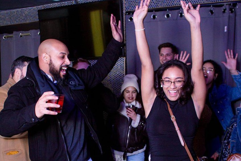 A group of people at a social event, some holding drinks, with raised hands and a person in the foreground wearing a black top. 2