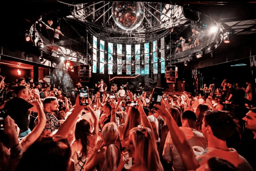 A crowded nightclub with a DJ booth, large screens, and a disco ball. 1