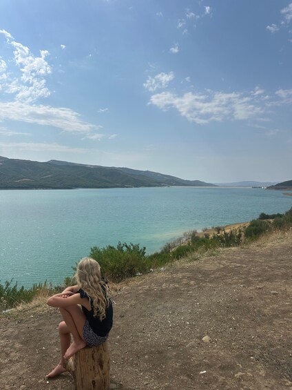 A person sitting on a wooden stump by a lake with mountains in the background. 2