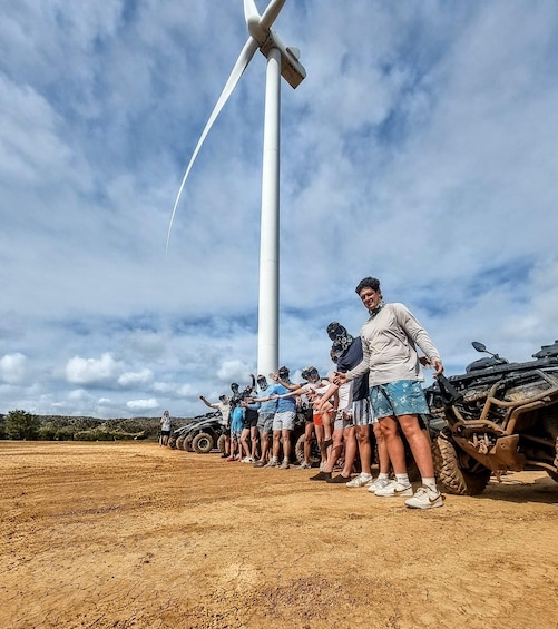 A group of people standing in a line with ATVs parked behind them, a large wind turbine in the background, and a dry, cracked ground. 2